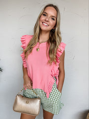 A woman smiling against a textured off-white wall, wearing a bubblegum pink sleeveless top crafted from a 50% viscose, 28% polyester, and 22% nylon knit blend. The top features a finely ribbed round crew neckline and dramatic ribbed ruffle trim framing the shoulders and extending down the open sides. Designed with an airy, open-side silhouette over a tan tube top, it is secured with matching knit side-tie closures at the waist. She is paired with green and white gingham patterned shorts, a gold toggle chain