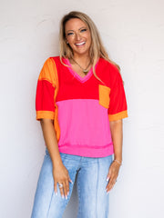 A portrait photograph focusing from the hips up of a smiling woman with long blonde wavy hair posing cheerfully against a textured white wall. She is wearing a short-sleeved colorblocked top featuring panels of bright red, hot pink, and orange made from an 80% cotton and 20% polyester blend fabric, designed with a distressed V-neckline, raw seam detailing, and an orange chest pocket, styled with light-wash jeans with front center seams, a gold heart pendant necklace, and stacked rings.
