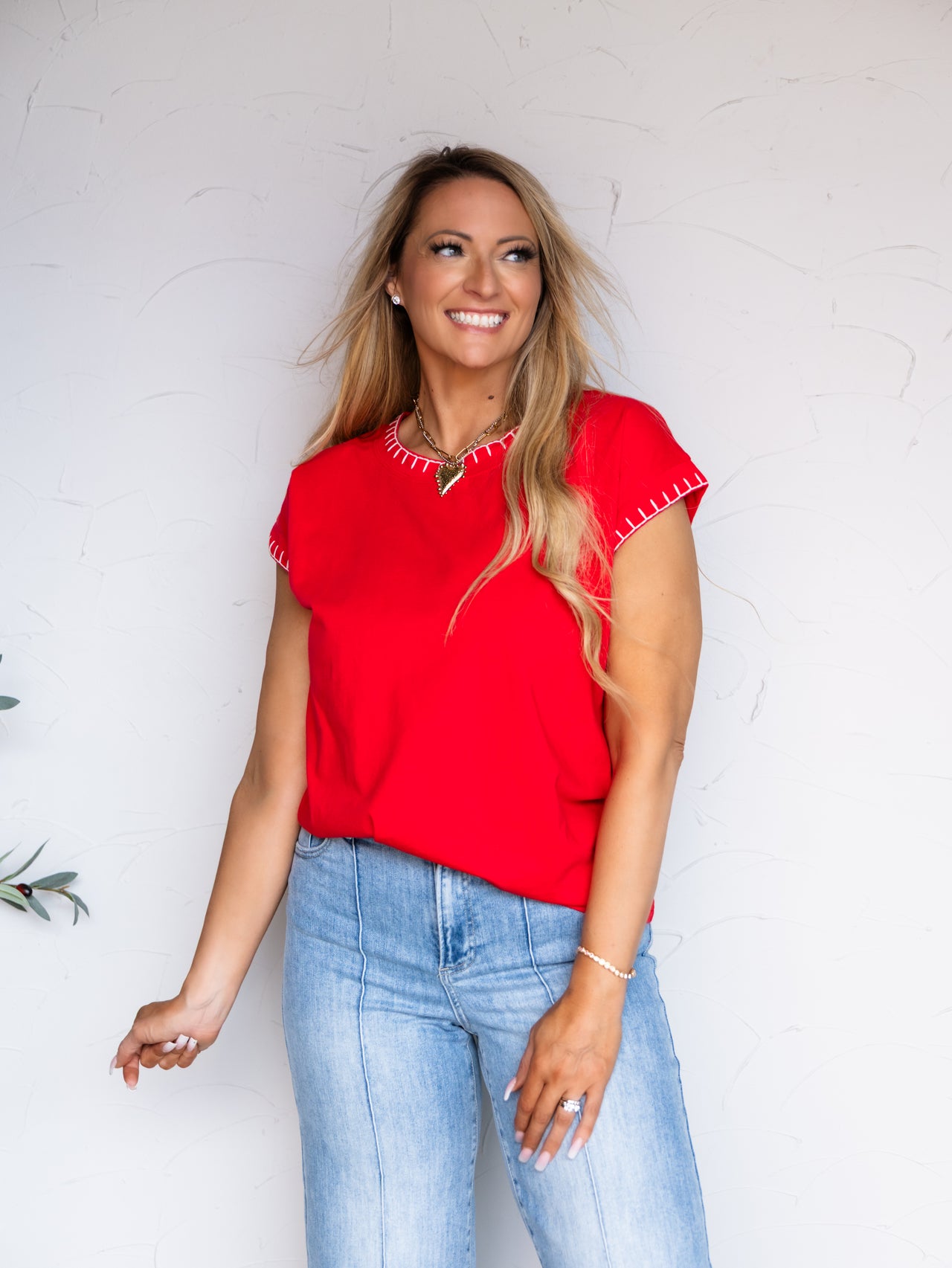 A smiling blonde woman wearing a bright red short-sleeve top that is 100% Cotton with decorative white stitching along the neckline and cuffs, paired with light blue jeans, standing against a white textured wall.