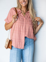 A portrait photograph focusing from the waist up of a smiling woman with long blonde wavy hair, glasses resting on top of her head, touching her hair while holding a purse strap against a textured white wall. She is wearing a dusty rose short-sleeved button-down blouse featuring subtle plaid grid texturing made from 100% cotton fabric, designed with a spread collar, cuffed dolman sleeves, and a chest pocket, styled with light-wash jeans with front center seams, a cognac brown and woven straw chain purse, an
