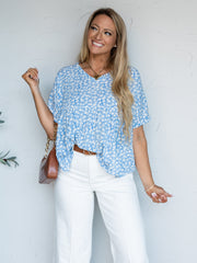 A portrait photograph focusing from the hips up of a smiling woman with long blonde wavy hair looking off to the side while holding a purse shoulder strap against a textured white wall. She is wearing a light blue short-sleeved blouse adorned with a delicate white floral print made from 100% polyester fabric, featuring a V-neckline, front tuck, and short dolman sleeves, styled with high-waisted white jeans, a cognac brown leather belt, a brown faux-croc chain bag, a gold heart necklace, and a bracelet.