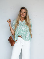 A portrait photograph focusing from the hips up of a smiling woman with long blonde wavy hair posing while holding a purse shoulder strap against a textured white wall. She is wearing a sage green short-sleeved blouse adorned with a delicate white floral print made from 100% polyester fabric, featuring a V-neckline, front tuck, and short dolman sleeves, styled with high-waisted white jeans, a cognac brown leather belt, a brown faux-croc chain bag, a gold heart necklace, and a bracelet.