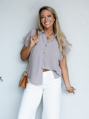 A portrait photograph focusing from the hips up of a smiling woman with long blonde wavy hair holding her purse strap against a textured white wall. She is wearing a soft taupe-colored short-sleeved button-down blouse made from 100% polyester fabric with a subtle crinkle texture, featuring a spread collar, cuffed dolman sleeves, and a single chest pocket, styled with high-waisted white jeans with front seams, a cognac brown leather belt, a brown and woven straw chain purse, and gold jewelry.