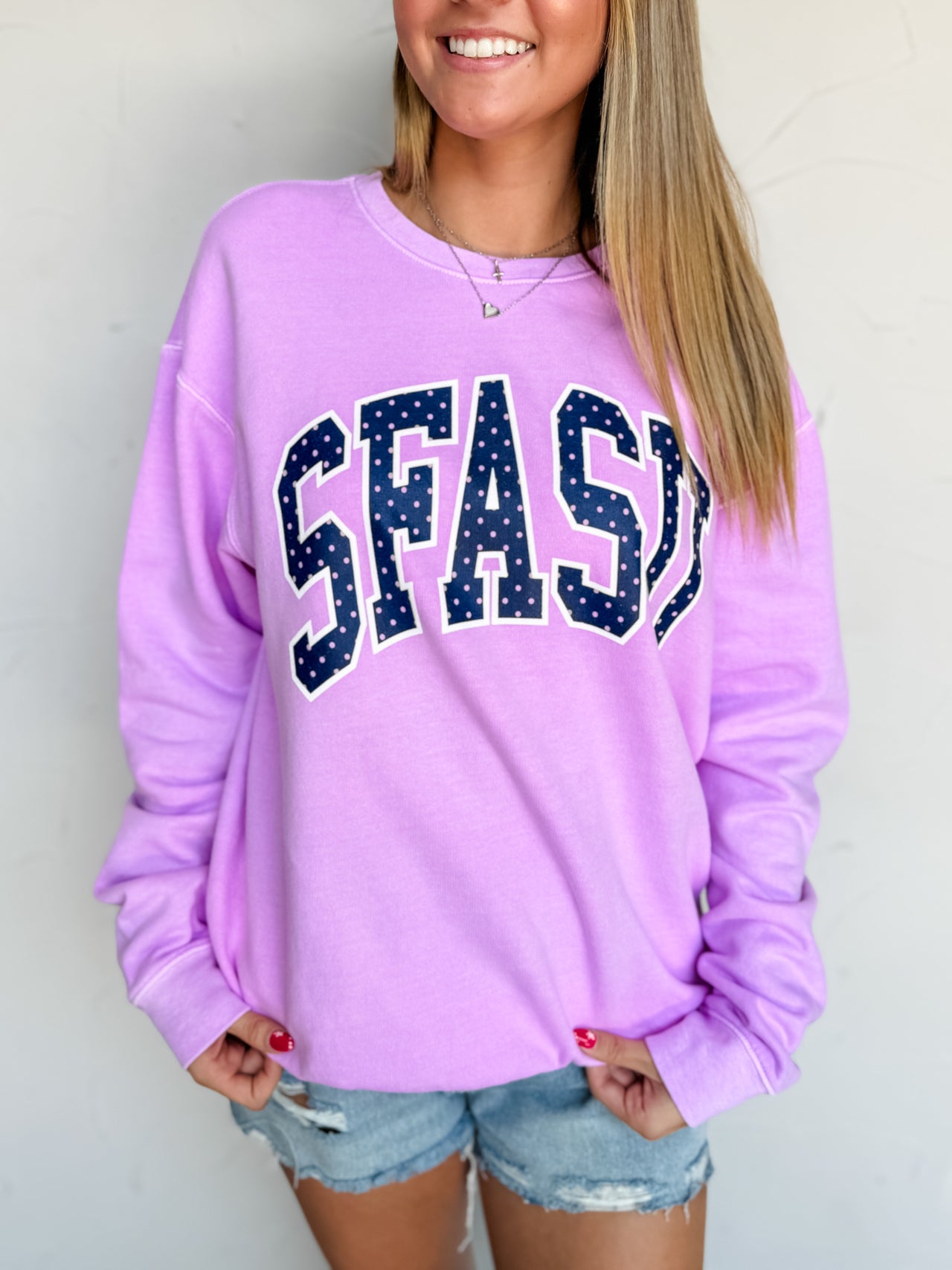 A woman wearing a 100% cotton lavender purple sweatshirt with 'SFASD' printed across the chest in navy blue letters with white dots, paired with denim shorts.