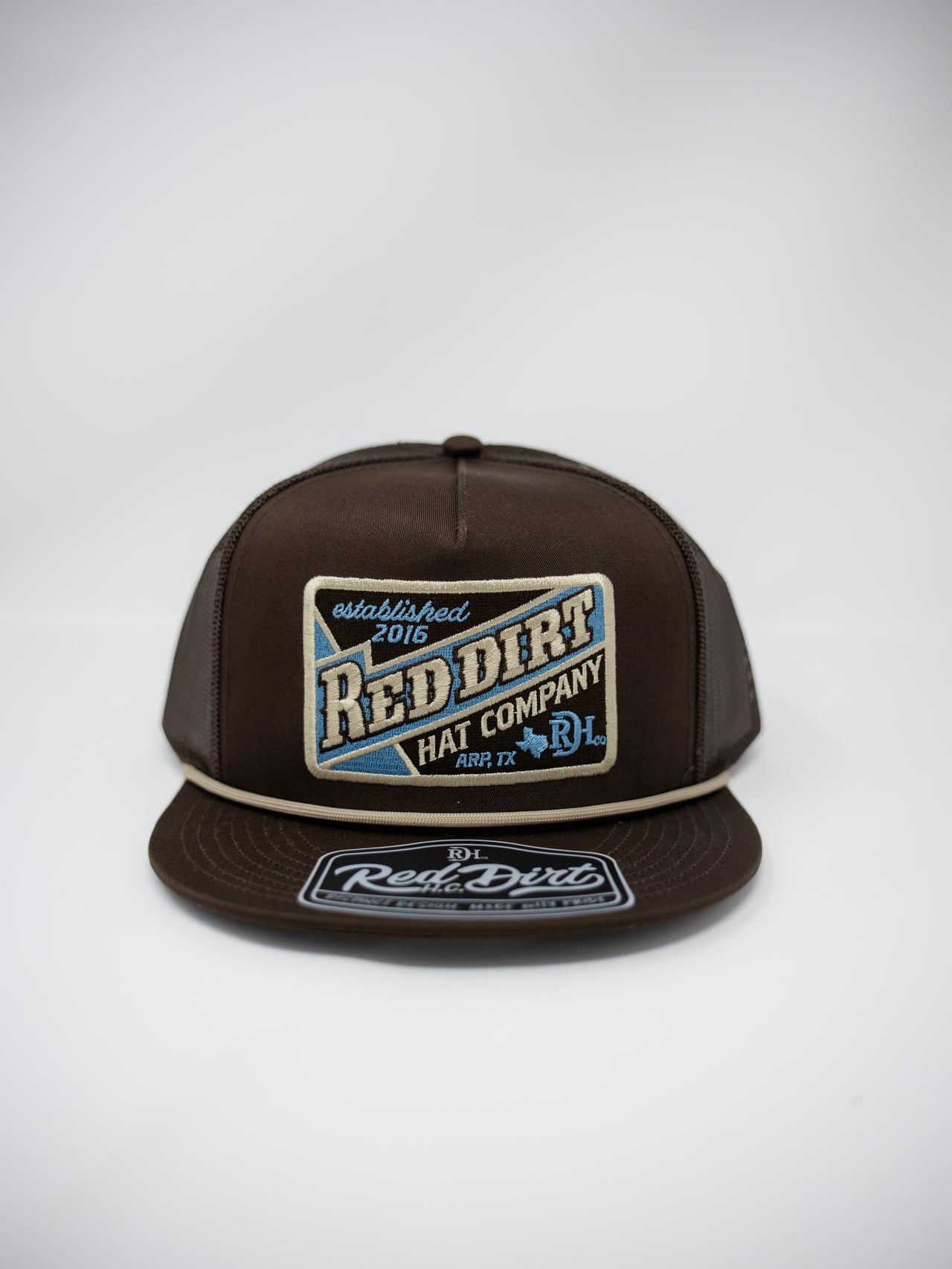 A dark brown snapback cap featuring a large embroidered front patch that reads 'Red Dirt Hat Company' with blue and cream details, and a matching embroidered visor patch.