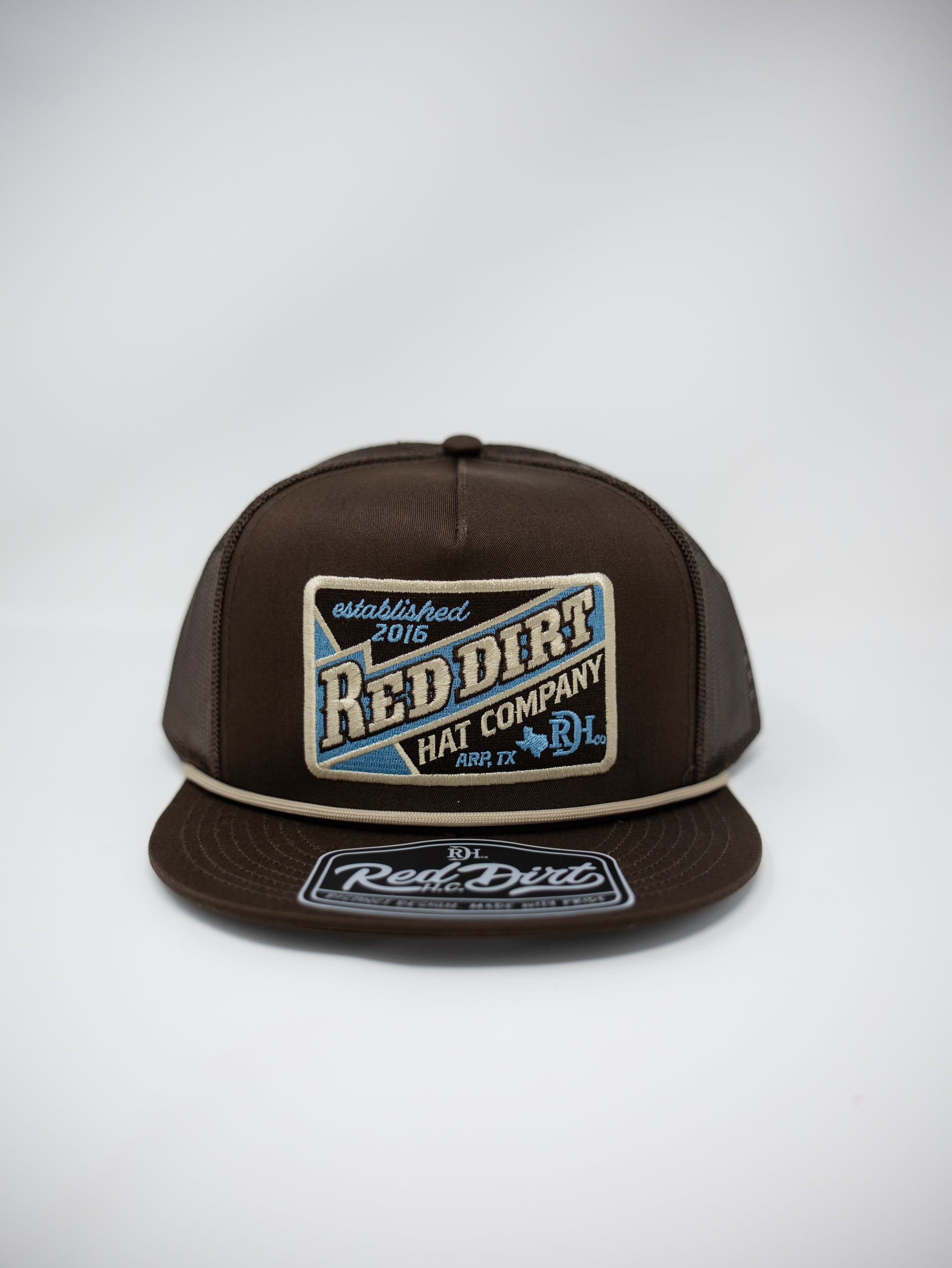 A dark brown snapback cap featuring a large embroidered front patch that reads 'Red Dirt Hat Company' with blue and cream details, and a matching embroidered visor patch.