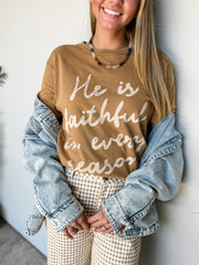 A woman wearing a 100% Cotton brown crewneck sweatshirt with white script text that reads 'He is faithful in every season', styled with a light-wash denim jacket draped off her shoulders and beige-and-white gingham pants.