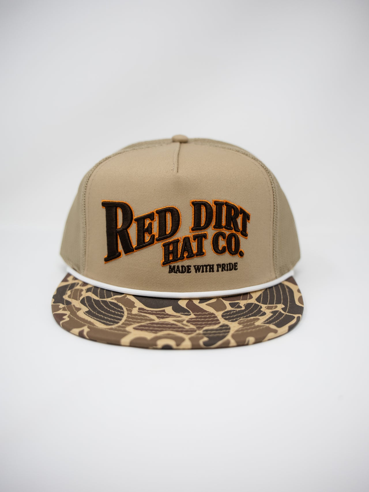 A tan snapback cap with 'RED DIRT HAT CO.' embroidered on the front in black with a brown outline, and a camouflage-patterned visor, shown against a light gray background.