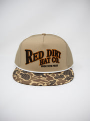A tan snapback cap with 'RED DIRT HAT CO.' embroidered on the front in black with a brown outline, and a camouflage-patterned visor, shown against a light gray background.