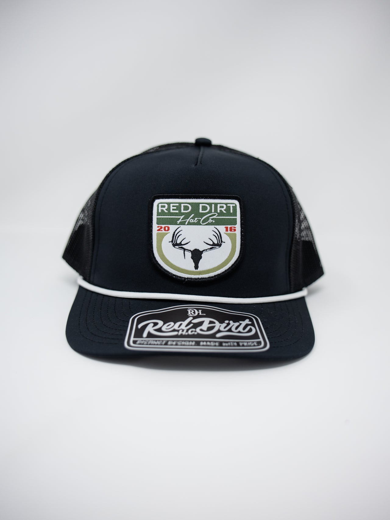 A front-facing shot of a black Red Dirt Hat Co. trucker cap with a white rope detail across the visor, set against a solid white background. The front panel features a patch with a buck skull silhouette, sage green accents, "2016" in red, and "RED DIRT Hat Co." text.