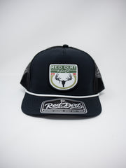 A front-facing shot of a black Red Dirt Hat Co. trucker cap with a white rope detail across the visor, set against a solid white background. The front panel features a patch with a buck skull silhouette, sage green accents, "2016" in red, and "RED DIRT Hat Co." text.