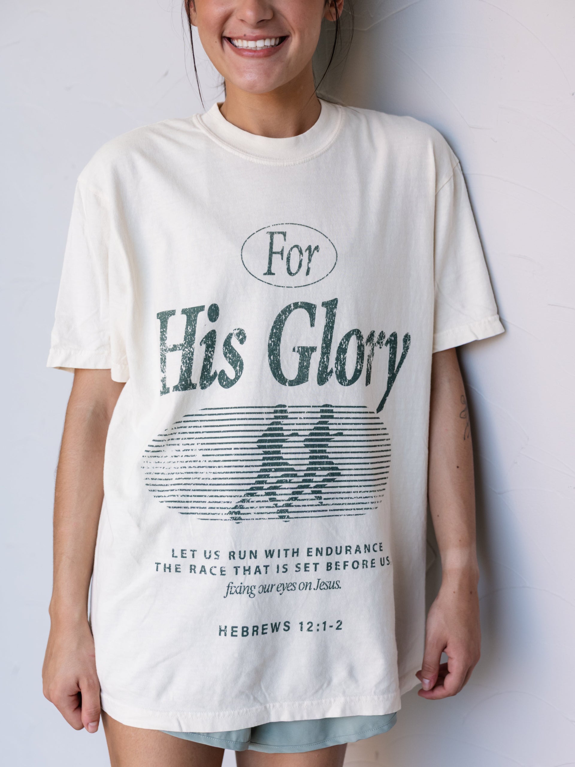 A woman wearing an oversized cream-colored graphic t-shirt with 'For His Glory' printed in dark green text and a running figure design, paired with light blue denim shorts, standing against a white wall.