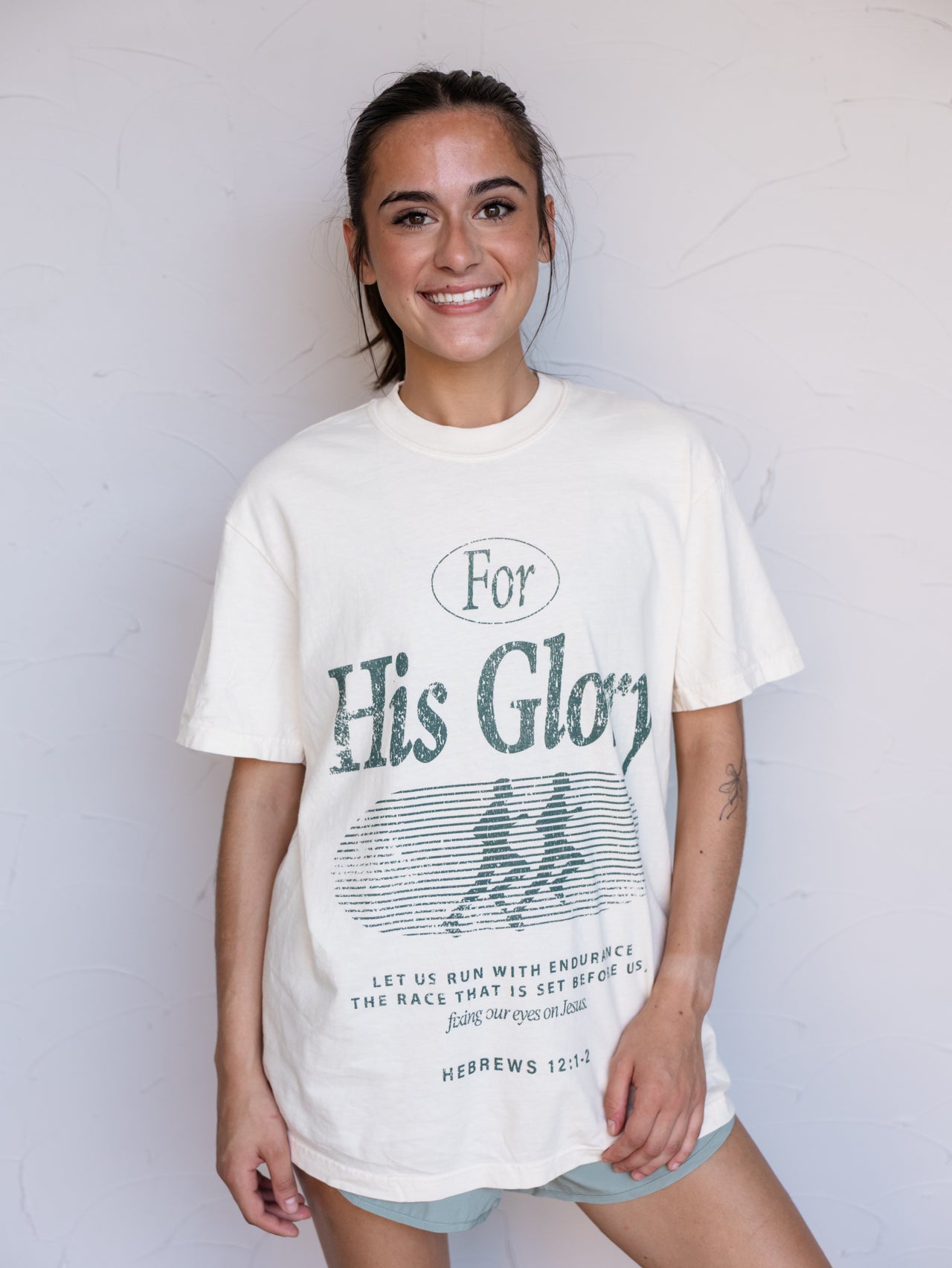 A woman wearing a cream-colored graphic t-shirt with a distressed green print that reads 'For His Glory' and 'Hebrews 12:2' on the front, standing against a white wall.