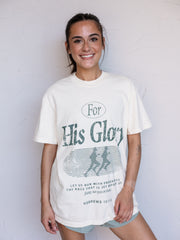 A woman wearing a cream-colored graphic t-shirt with a distressed green print that reads 'For His Glory' and 'Hebrews 12:2' on the front, standing against a white wall.