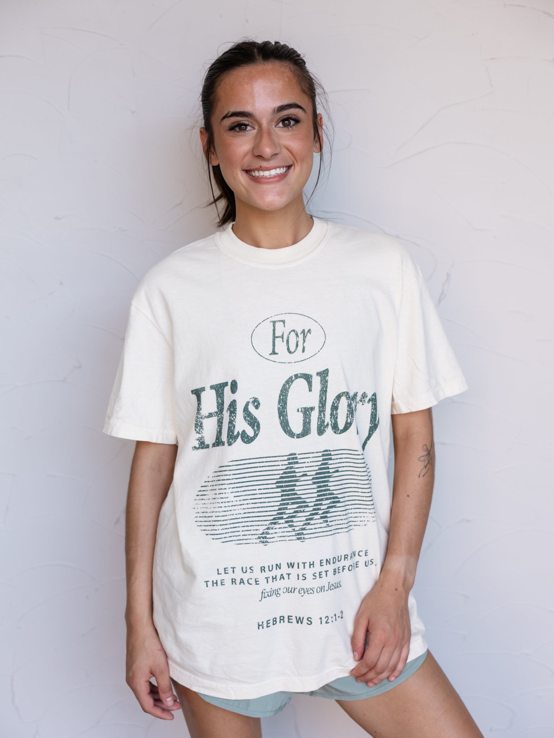 A woman wearing a cream-colored graphic t-shirt with a distressed green print that reads 'For His Glory' and 'Hebrews 12:2' on the front, standing against a white wall.