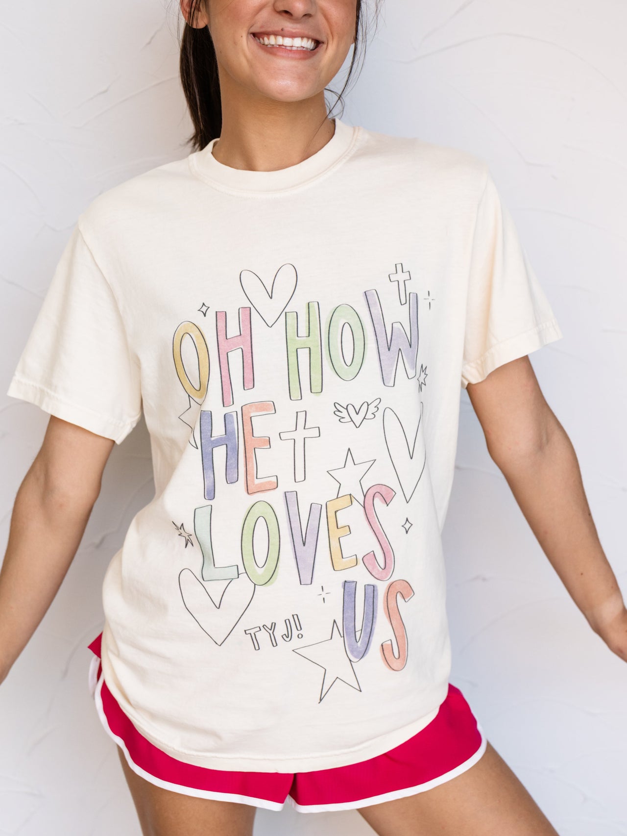 A woman wearing a cream-colored graphic t-shirt with pastel-colored text that reads 'OH HOW HE LOVES US' and red athletic shorts, posing against a white wall.