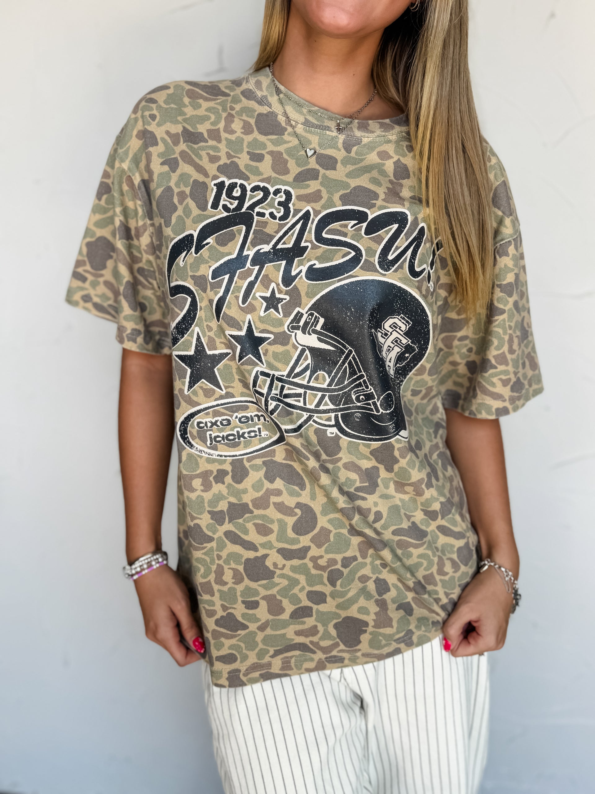 A woman wearing a 100% Cotton vintage-style camouflage graphic t-shirt with a football helmet and '1923 STASIS' print, paired with white pinstriped pants, standing against a white wall.