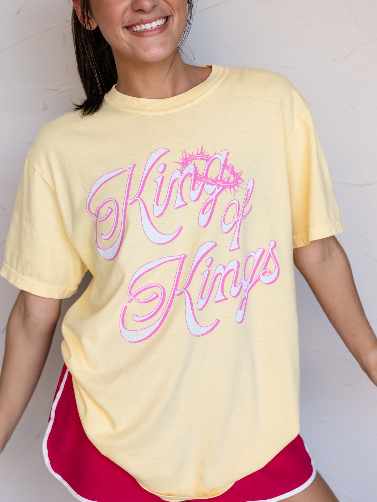 A woman wearing a pale yellow oversized graphic T-shirt with 'King of Kings' in pink and white script, paired with red athletic shorts, against a light textured wall.