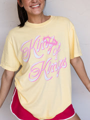 A woman wearing a pale yellow oversized graphic T-shirt with 'King of Kings' in pink and white script, paired with red athletic shorts, against a light textured wall.