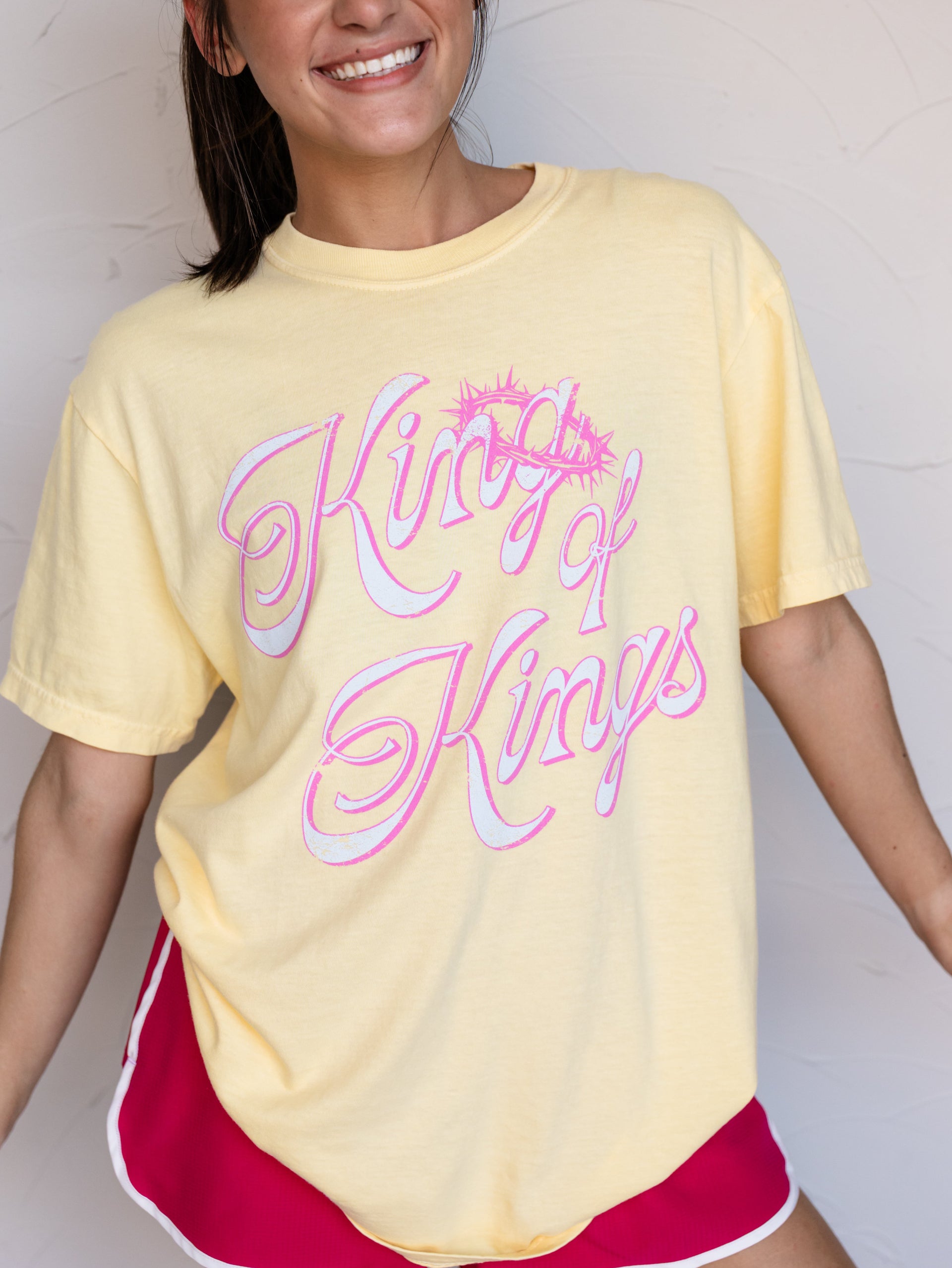 A woman wearing a pale yellow oversized graphic T-shirt with 'King of Kings' in pink and white script, paired with red athletic shorts, against a light textured wall.