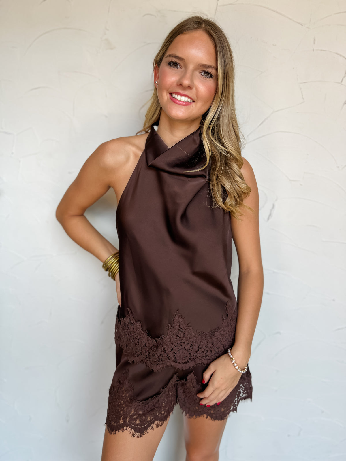 A waist-up photograph of a smiling blonde woman with wavy hair posing with one hand on her hip against a textured white wall. She is wearing a matching two-piece chocolate brown satin set made from 100% polyester, including a sleeveless high-neck halter top with a soft draped front and scalloped lace trim along the hem, paired with matching satin shorts featuring a lace-trimmed hem, styled with stacked gold bangles, a delicate beaded bracelet, and stud earrings.