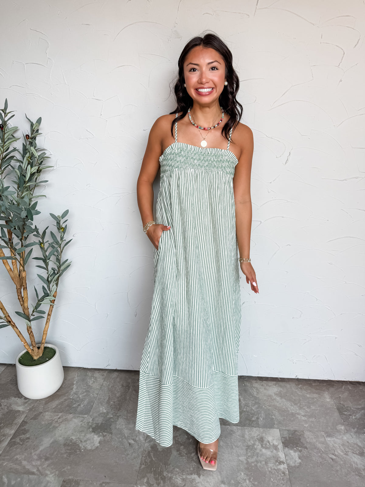 A full-length photograph of a smiling woman with dark wavy hair posing with one hand in her pocket against a textured white wall next to a potted olive plant. She is wearing a sleeveless sage green and white vertically striped maxi dress made from an 80% polyester, 16% rayon, and 2% spandex blend, featuring spaghetti straps, a smocked elastic stretch bust, functional side pockets, and a wide hem band, styled with clear block heels, a colorful beaded necklace, a gold medallion pendant, pearl earrings, and go