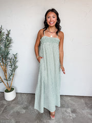 A full-length photograph of a smiling woman with dark wavy hair posing with one hand in her pocket against a textured white wall next to a potted olive plant. She is wearing a sleeveless sage green and white vertically striped maxi dress made from an 80% polyester, 16% rayon, and 2% spandex blend, featuring spaghetti straps, a smocked elastic stretch bust, functional side pockets, and a wide hem band, styled with clear block heels, a colorful beaded necklace, a gold medallion pendant, pearl earrings, and go