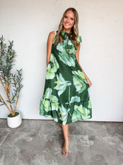 A full-length photograph of a smiling young woman with wavy brown hair posing against a textured white wall next to a potted olive plant. She is wearing a sleeveless emerald green midi dress made from 100% polyester, featuring a large-scale watercolor floral print in shades of mint and lime green, a ruffled high collar with a tie bow, and a tiered ruffle hem, styled with clear strap slide heels and delicate jewelry.
