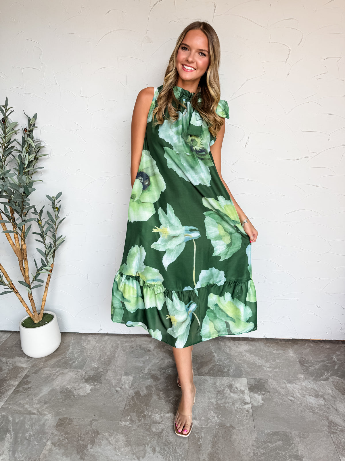 A full-length photograph of a smiling young woman with wavy brown hair posing against a textured white wall next to a potted olive plant. She is wearing a sleeveless emerald green midi dress made from 100% polyester, featuring a large-scale watercolor floral print in shades of mint and lime green, a ruffled high collar with a tie bow, and a tiered ruffle hem, styled with clear strap slide heels and delicate jewelry.
