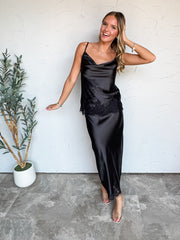 A full-length photograph of a smiling blonde woman with wavy hair touching her head while posing against a textured white wall next to a potted olive plant. She is wearing a matching two-piece black satin set made from 100% polyester, featuring a sleeveless cowl-neck Cami top with a black lace trim hemline and a high-waisted bias-cut satin midi skirt with matching lace trim along the hem, styled with clear strap heels, delicate silver necklaces, and silver chain bracelets.