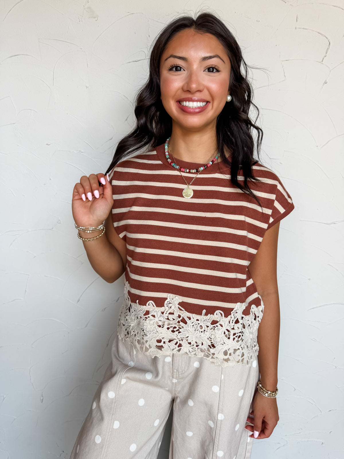A waist-up photograph of a smiling woman with dark wavy hair touching a section of her hair while posing against a textured white wall. She is wearing a short-sleeved fine-knit top made from a 50% viscose, 25% polyester, and 25% nylon blend, featuring horizontal rust brown and cream stripes with a scalloped ivory crochet lace trim along the hem, paired with beige polka dot pants and styled with a colorful beaded necklace, a gold medallion pendant, pearl stud earrings, and stacked gold bracelets.