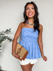 A close-up, front-facing photo cropped at the chin of a woman standing against a textured white wall, wearing a strapless blue and white gingham peplum top. The top features a structured, fitted bodice that flows into a sharply pleated, flared peplum waist. She is accessorized with a colorful beaded necklace featuring shades of blue, green, and gold, stacked gold bracelets, and a light blue manicure. Under her arm.