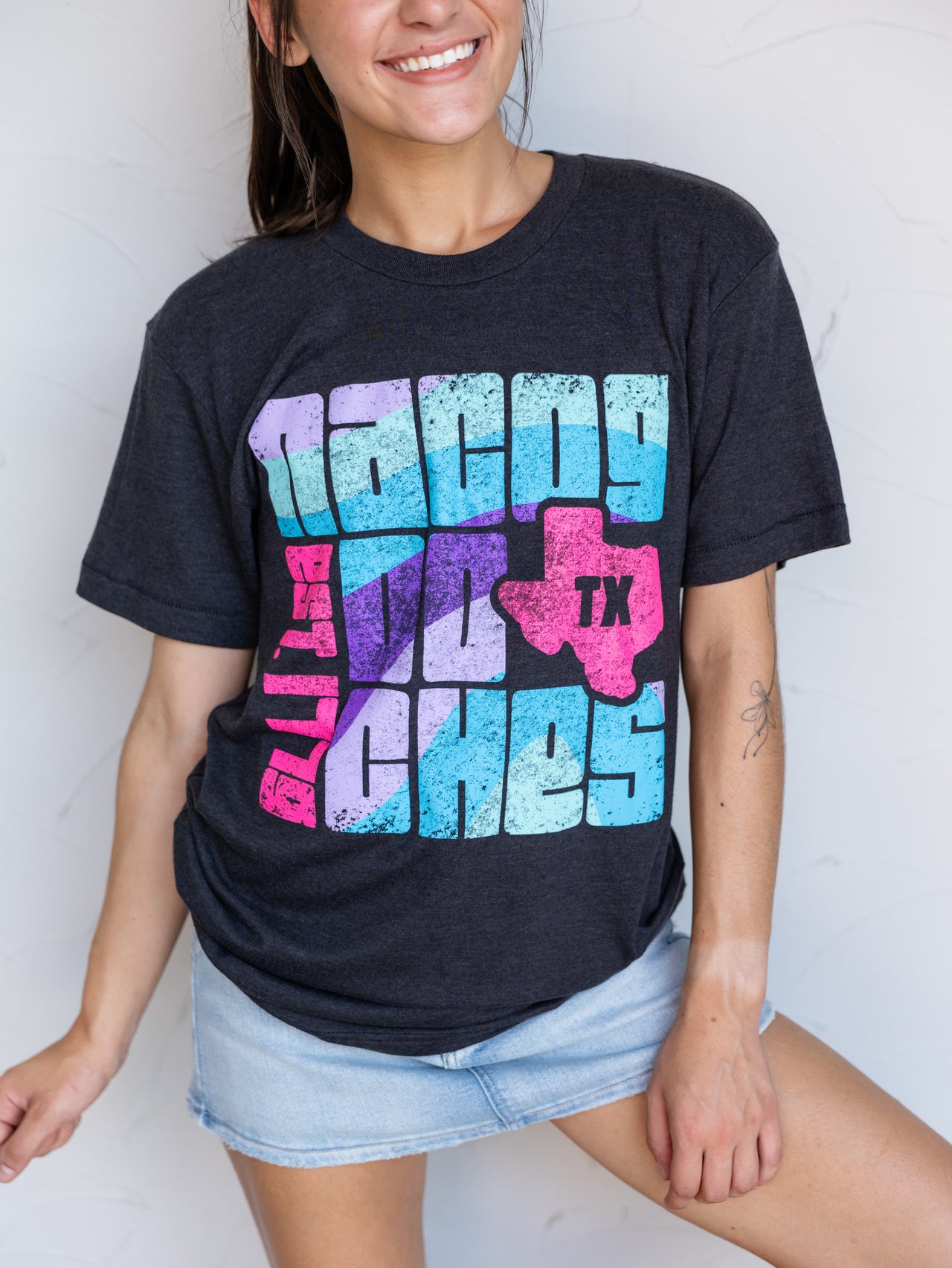 A woman wearing a dark charcoal heather graphic t-shirt with a colorful distressed print that reads 'Nacyo and the Zones' with a pink Texas-shaped outline containing 'TX'. She is wearing light-wash denim shorts against a white wall.