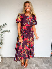 A full-length photograph of a smiling blonde woman with long curly hair posing against a textured off-white wall next to a potted olive tree. She is wearing a short-sleeved maxi dress made from an 84% viscose and 16% nylon blend, featuring a deep plum purple background with vibrant coral pink, orange, and blue floral patterns, a high crew neckline, elasticized short puff sleeves, an elastic waist, and a multi-tiered ruffle skirt, styled with cognac brown leather slide sandals, a gold chain necklace, and sta