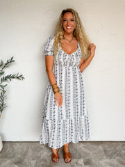 A three-quarter length photograph of a smiling blonde woman with long curly hair touching her strands while posing with one hand in her pocket against a textured white wall next to an olive branch. She is wearing a short-sleeved deep plum purple fine-knit top made from a 60% polyester and 40% viscose blend, featuring a wide vertical ribbed texture, crew neckline, and elbow-length sleeves, front-tucked into magenta pants with pink oversized polka dots, styled with sunglasses resting on her head, a gold chain