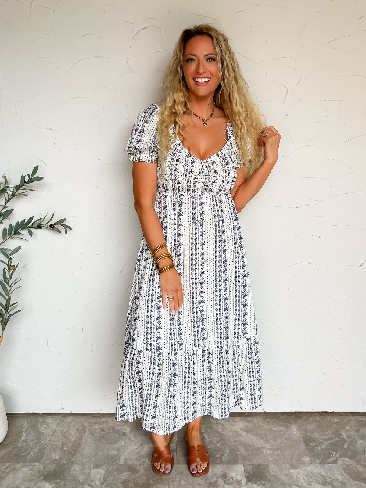 A three-quarter length photograph of a smiling blonde woman with long curly hair touching her strands while posing with one hand in her pocket against a textured white wall next to an olive branch. She is wearing a short-sleeved deep plum purple fine-knit top made from a 60% polyester and 40% viscose blend, featuring a wide vertical ribbed texture, crew neckline, and elbow-length sleeves, front-tucked into magenta pants with pink oversized polka dots, styled with sunglasses resting on her head, a gold chain