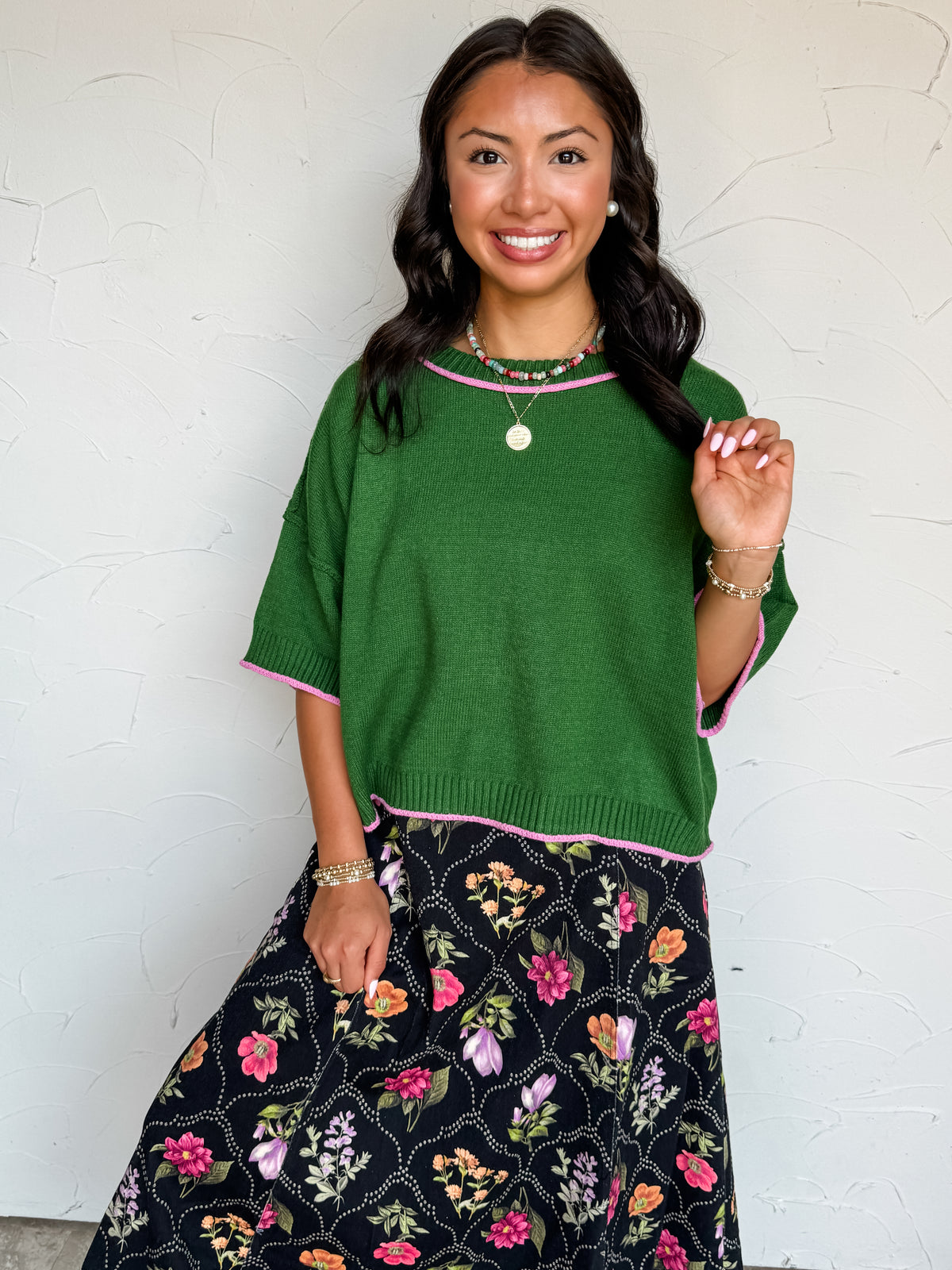 A waist-up photograph of a smiling woman with dark wavy hair touching a section of her hair while posing against a textured white wall. She is wearing a short-sleeved fine-knit forest green sweater top made from a 50% viscose, 28% polyester, and 22% nylon blend, featuring bubblegum pink contrast stitch trim along the crew neckline, sleeve cuffs, and bottom hem, paired with a black floral printed midi skirt and accessorized with a colorful beaded necklace, a gold medallion pendant, pearl earrings, and stacke
