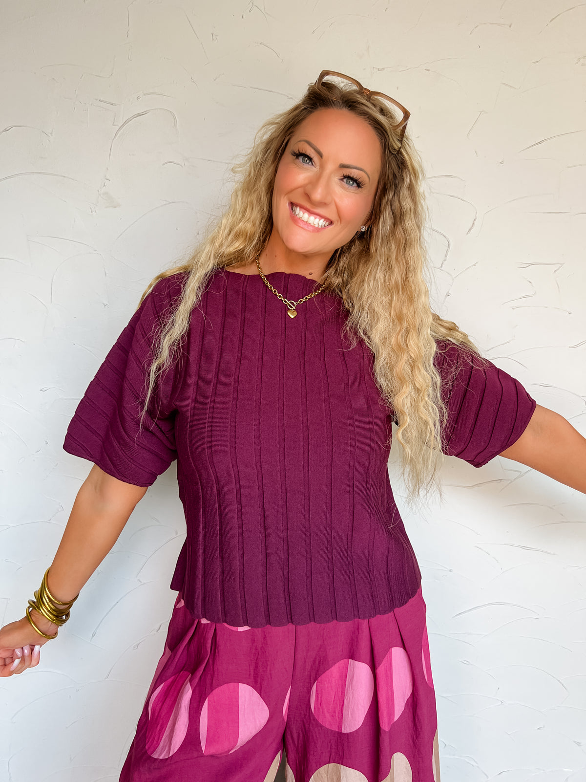 A three-quarter length photograph of a smiling blonde woman with long curly hair touching her strands while posing with one hand in her pocket against a textured white wall next to an olive branch. She is wearing a short-sleeved deep plum purple fine-knit top made from a 60% polyester and 40% viscose blend, featuring a wide vertical ribbed texture, crew neckline, and elbow-length sleeves, front-tucked into magenta pants with pink oversized polka dots, styled with sunglasses resting on her head, a gold chain