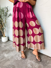A photograph from the waist down of a model leaning against a textured white wall next to a potted olive plant in a studio setting. She is wearing high-waisted, wide-leg pants made from an 85% rayon and 15% nylon blend, featuring a rich magenta base with oversized polka dots divided into dual-tone shading in shades of pink, beige, and tan, styled with a gathered elastic waistband, front pleat details, flat metallic slide sandals, and a plum purple top.