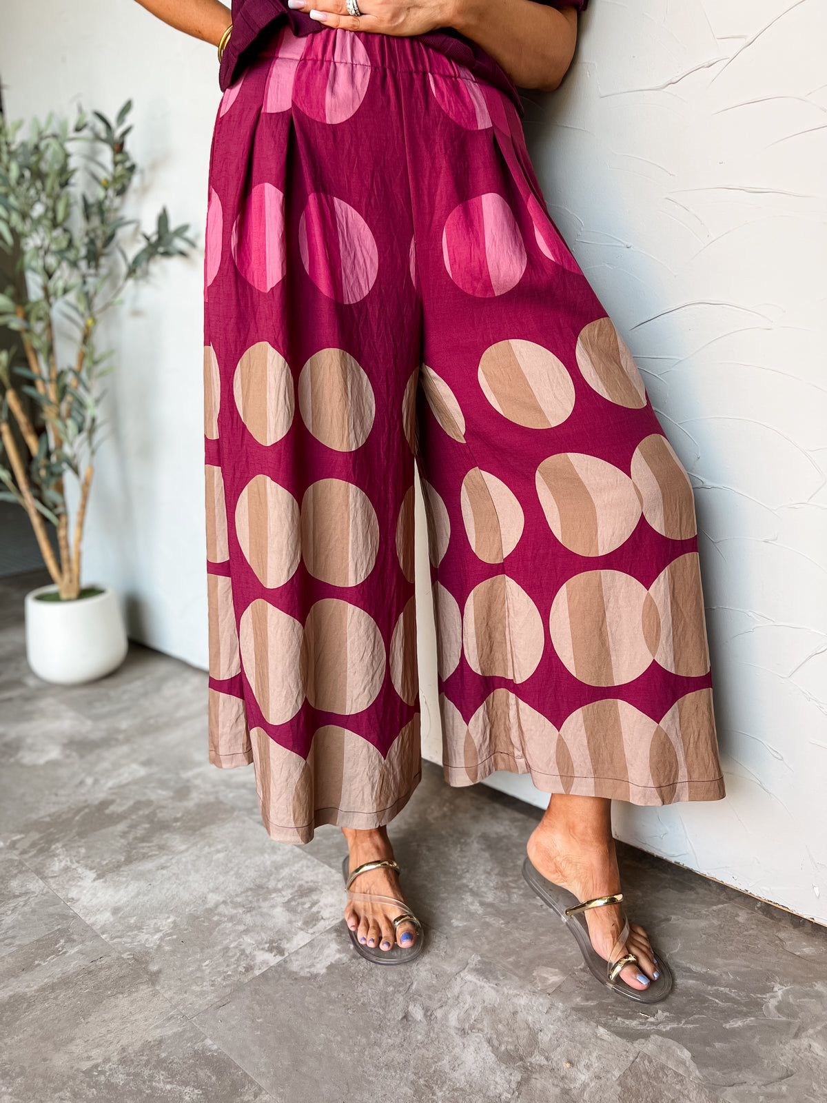A photograph from the waist down of a model leaning against a textured white wall next to a potted olive plant in a studio setting. She is wearing high-waisted, wide-leg pants made from an 85% rayon and 15% nylon blend, featuring a rich magenta base with oversized polka dots divided into dual-tone shading in shades of pink, beige, and tan, styled with a gathered elastic waistband, front pleat details, flat metallic slide sandals, and a plum purple top.
