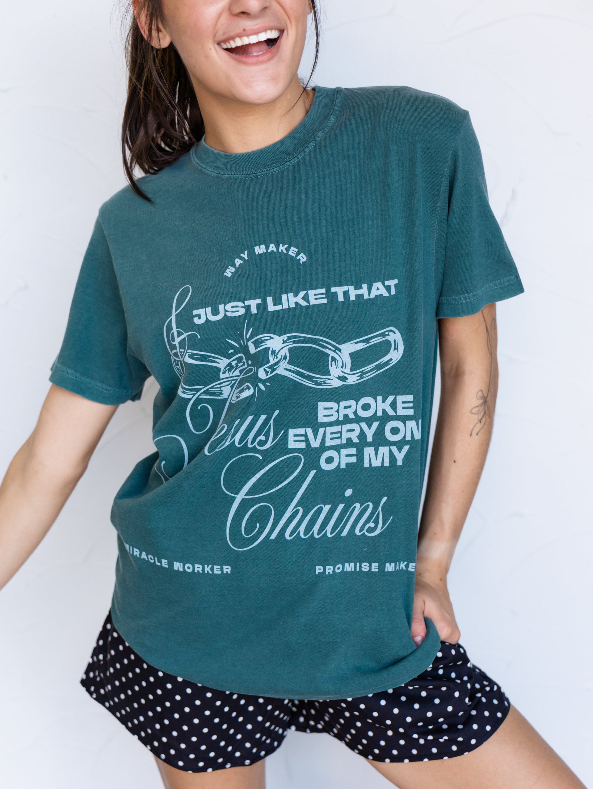 A woman wearing a teal graphic t-shirt with white text and chain illustrations, paired with black and white polka dot shorts, posing against a white wall.