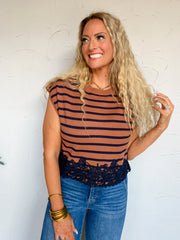 A three-quarter length photograph of a smiling blonde woman with long curly hair touching her strands while posing against a textured off-white wall. She is wearing a sleeveless fine-knit top made from a 50% viscose, 25% polyester, and 25% nylon blend, featuring horizontal camel brown and dark navy stripes, a crew neckline, cap shoulders, and a detailed dark navy lace scalloped hemline, paired with medium-wash blue denim jeans, styled with a gold chain necklace and stacked gold bangles.