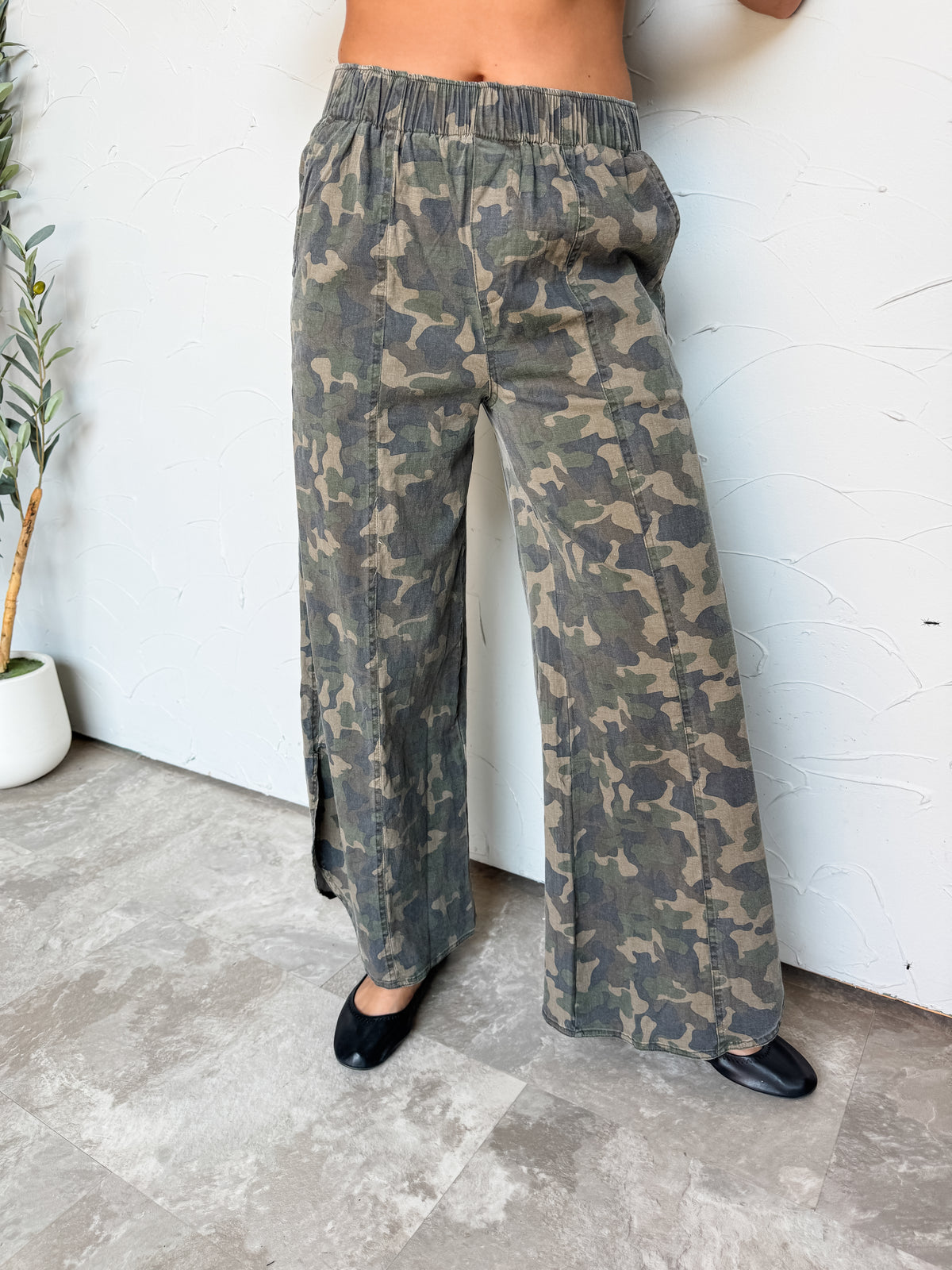 A lower-body photograph of a woman standing against a textured white wall, posing in wide-leg pants. She is wearing high-waisted olive green camouflage pants made from a 97% cotton and 3% spandex blend, featuring a smocked elastic waistband, vertical center leg seams, side pockets, and small side slits at the hem, styled with black ballet flats.