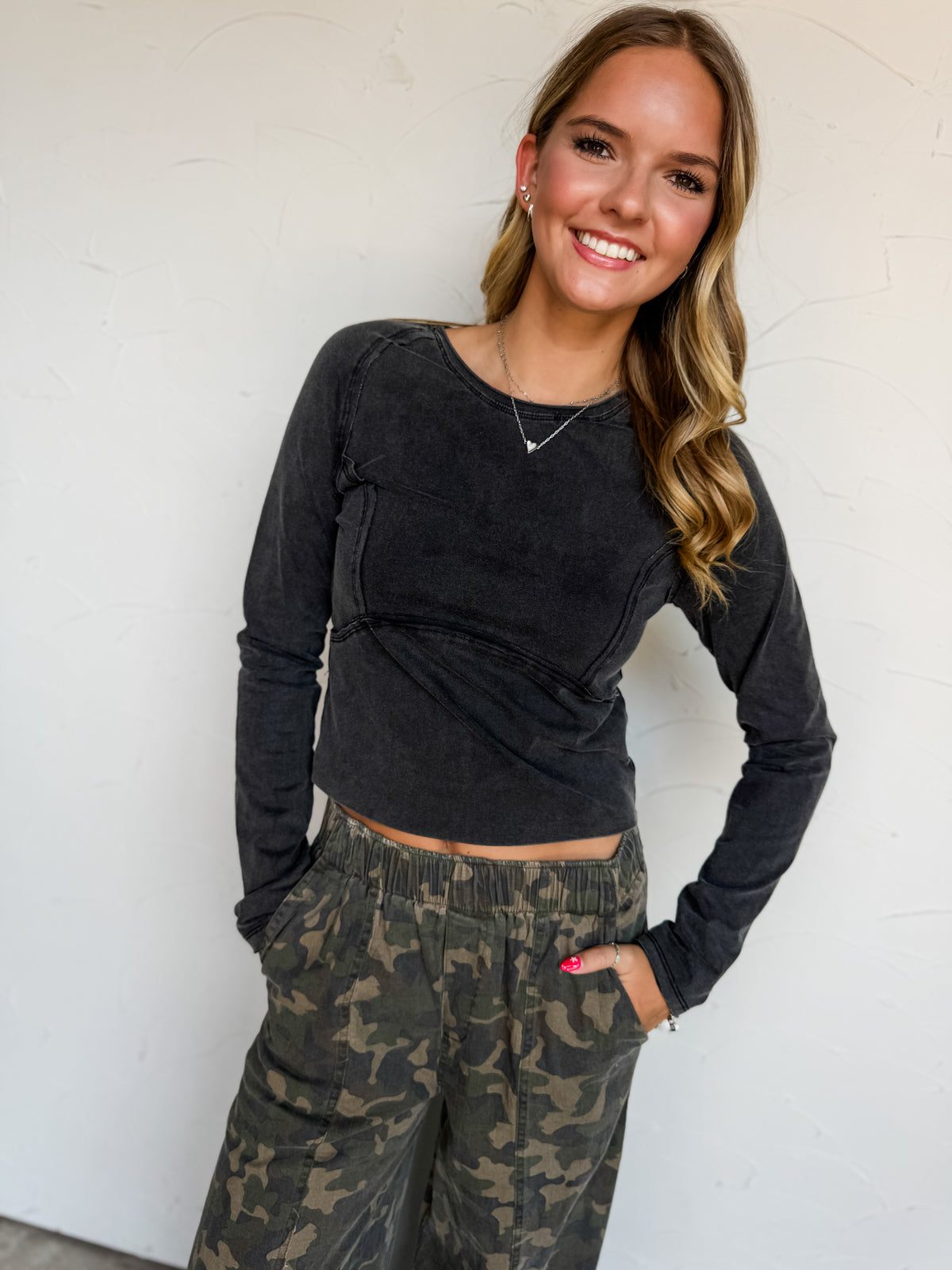 A waist-up photograph of a smiling young blonde woman posing with her hands in her pockets against a textured white wall. She is wearing a dark washed black long-sleeved top made from a 95% cotton and 5% spandex blend, featuring a crew neckline and exposed seam detailing across the chest and raglan shoulders, paired with camouflage wide-leg pants and styled with a silver heart pendant necklace, stud earrings, and silver bracelets.