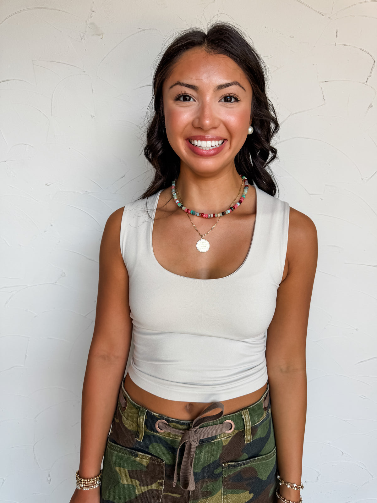 A waist-up photograph of a smiling woman with dark wavy hair posing against a textured white wall. She is wearing a fitted off-white tank top made from a 90% nylon and 10% spandex blend, featuring a scoop neckline and thick shoulder straps, paired with tie-front camouflage pants, and styled with a colorful beaded necklace, a gold medallion pendant, pearl stud earrings, and stacked gold beaded bracelets.
