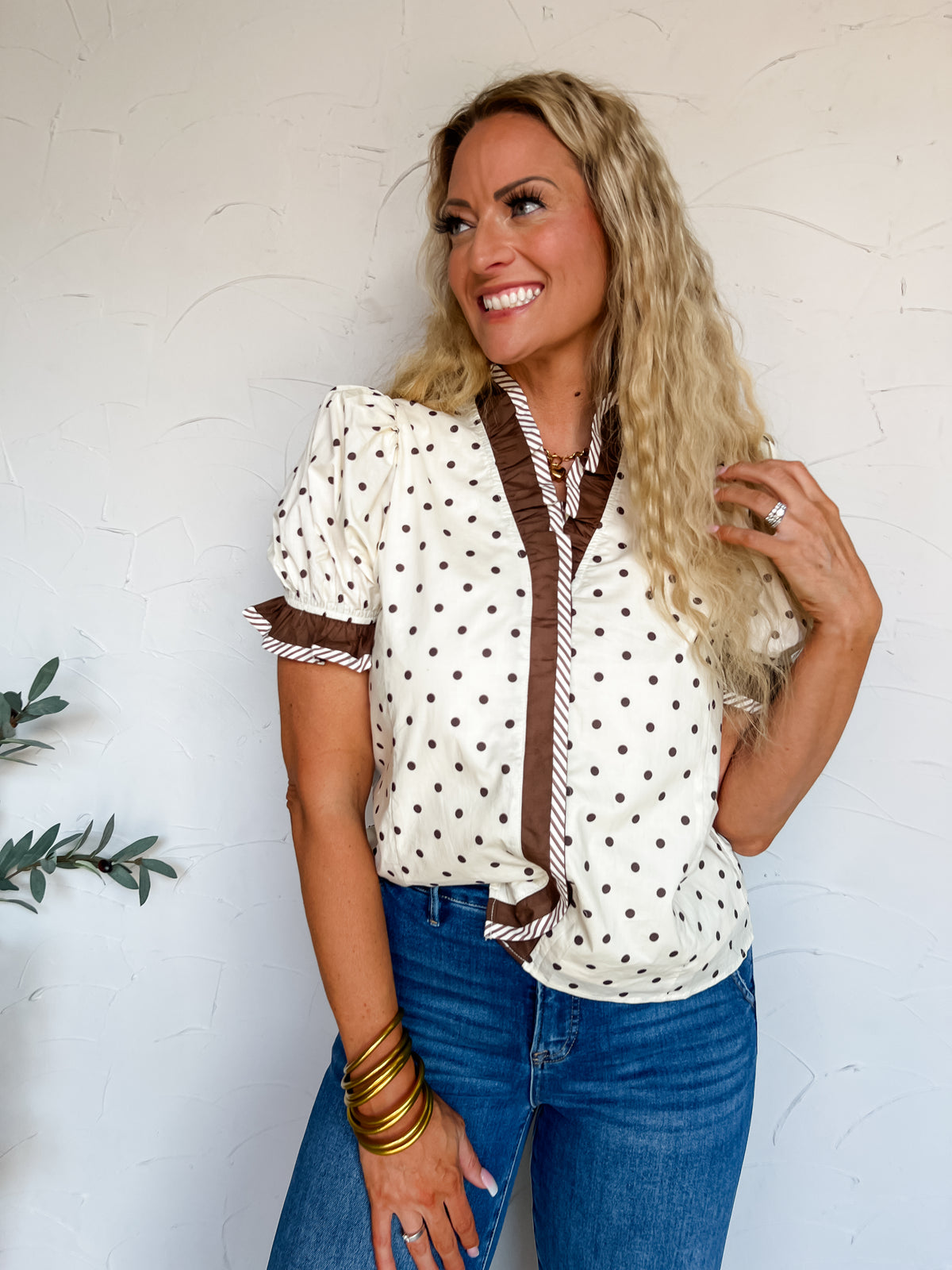 A three-quarter length photograph of a smiling blonde woman with long curly hair touching her hair while posing against a textured white wall next to an olive branch. She is wearing a short-sleeved 100% cotton cream blouse with chocolate brown polka dots, featuring a V-neckline trimmed with a chocolate brown ruffle with brown and white diagonal stripe edging, puff sleeves with matching ruffled cuffs, tucked into medium-wash blue denim jeans, styled with stacked gold bangles and a gold chain necklace.