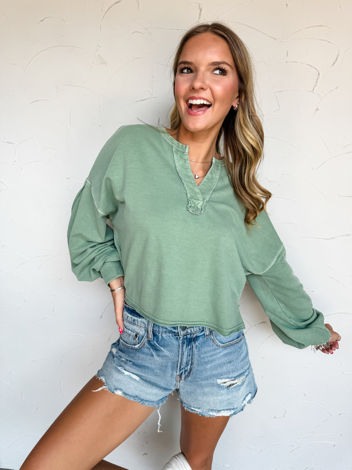 A waist-up photograph of an excited blonde woman with subtle waves looking off-camera with an open-mouth smile against a textured white wall. She is wearing a soft sage green long-sleeved pullover sweatshirt made from a 60% cotton and 40% polyester blend, featuring a split V-neckline with raw-edge trim, dropped shoulders, and wide balloon sleeves, paired with distressed light-wash denim shorts and styled with silver hoop earrings, a delicate heart pendant necklace, and stacked silver bracelets.