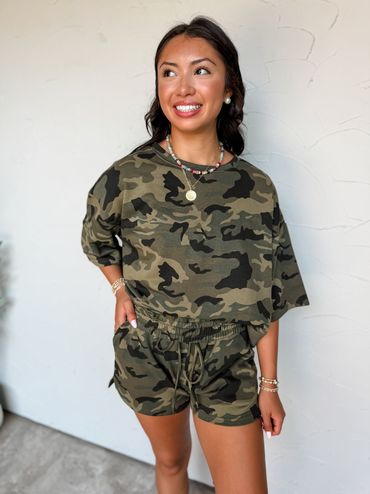 A waist-up photograph of a smiling dark-haired woman posing with one hand in her pocket against a textured white wall. She is wearing a matching two-piece camouflage lounge set made from a 60% cotton and 40% polyester blend, featuring an oversized short-sleeved crewneck tee and pull-on shorts with an elastic drawstring waistband, styled with a colorful beaded necklace, a gold medallion pendant, pearl earrings, and beaded bracelets.