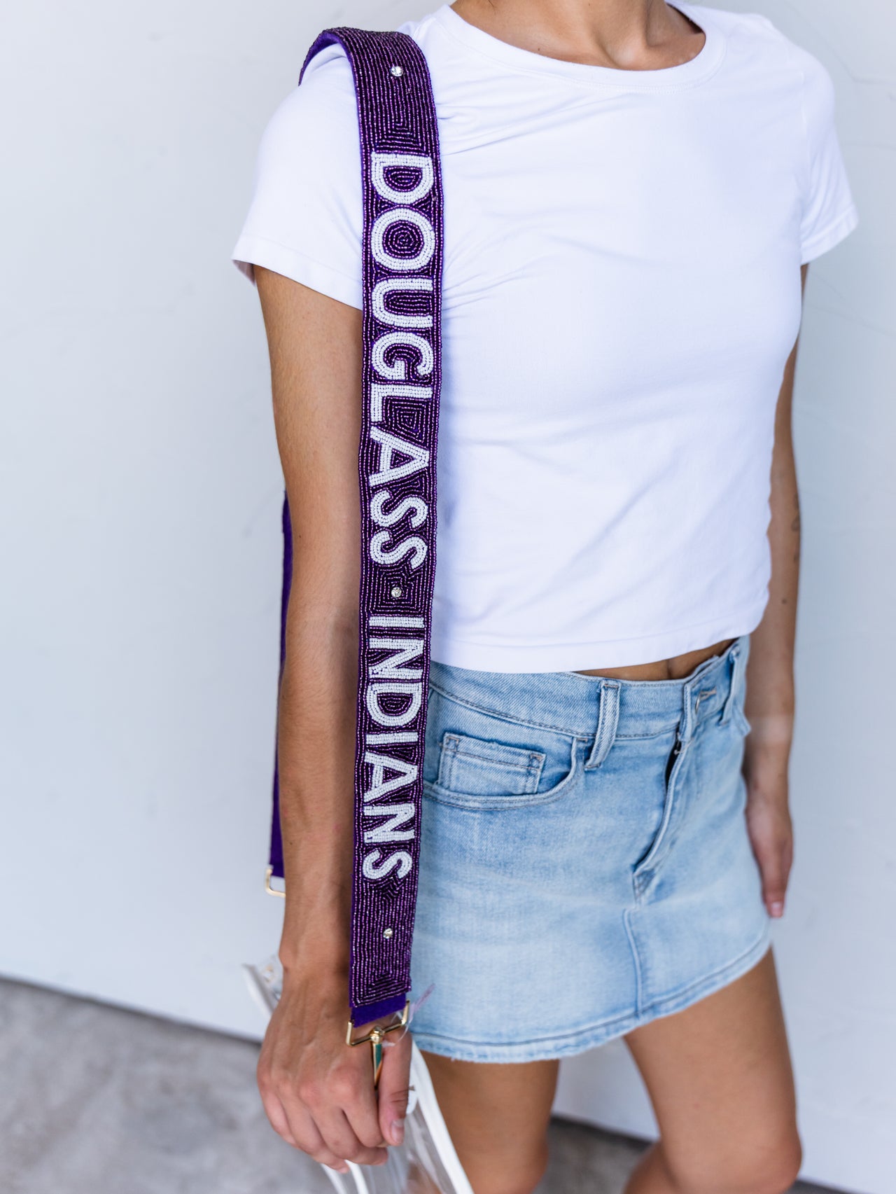 A person wearing a white t-shirt and denim skirt carries a purple beaded shoulder strap with 'DOUGLAS INDIANS' printed in white. The product is the shoulder strap, shown worn over the shoulder.