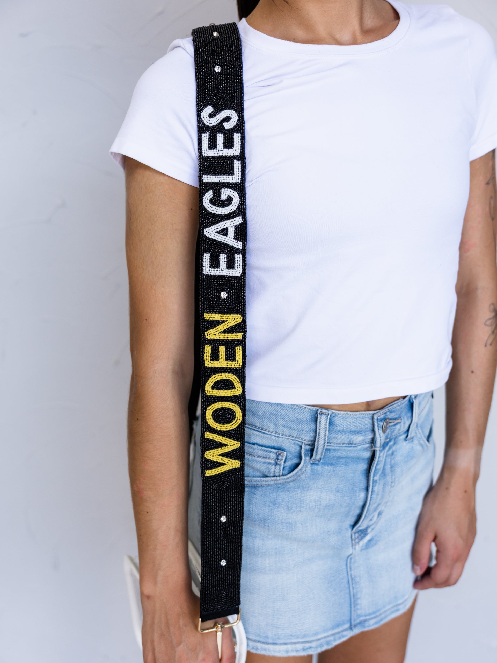 A close-up of a person wearing a white t-shirt and denim skirt, featuring a black crossbody bag strap with 'WODEN • EAGLES' text in white and yellow.